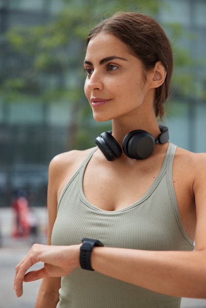 Free photo woman dressed in activewear checks results of fitness training wears wireless headphones around neck poses on blurred