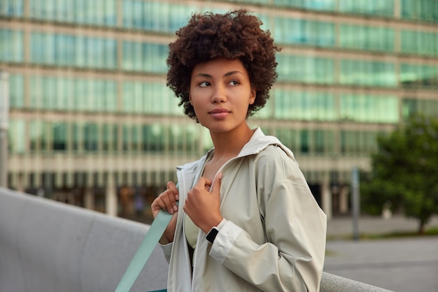 woman dressed in jacket carries karemat being on her way to sport studio looks away thoughtfully poses outdoors