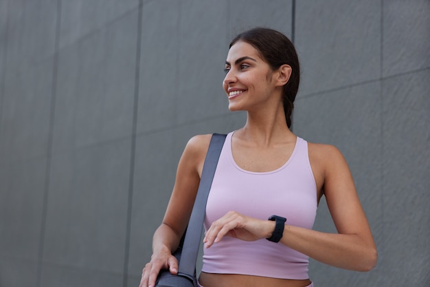  woman dressed in sportswear wears smartwatch carries rolled karemat looks away with gentle smile poses near grey wall being in good mood