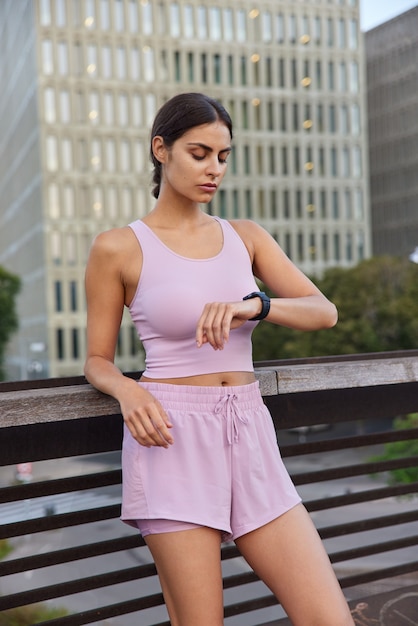  woman dressed in tank top and shorts checks time on smartwatch poses on bridge on modern city building waits someone to start training together. Fitness routine for health