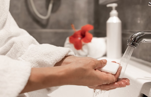 A woman in a dressing gown washes her hands with soap under running water from a tap.