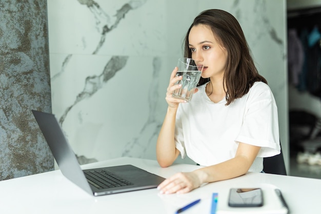 Woman Drinking from Water Glass while Typing at her Laptop in staying hydrated while working from home