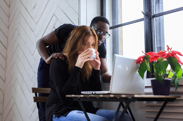 A woman drinking morning coffee and a black manin eyeglasses using a laptop.