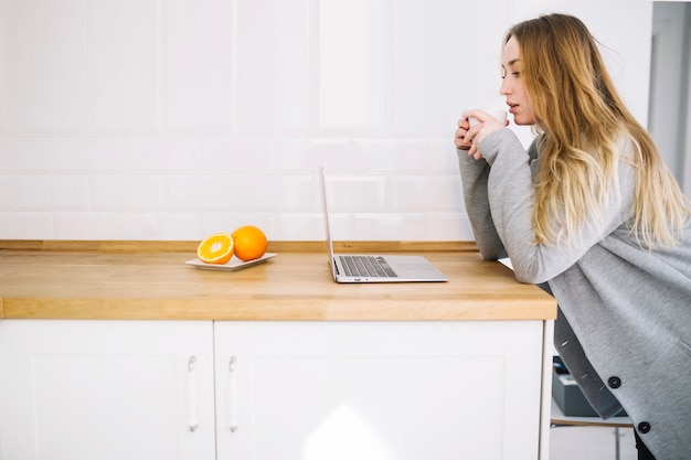 Free photo woman drinking and using laptop in kitchen