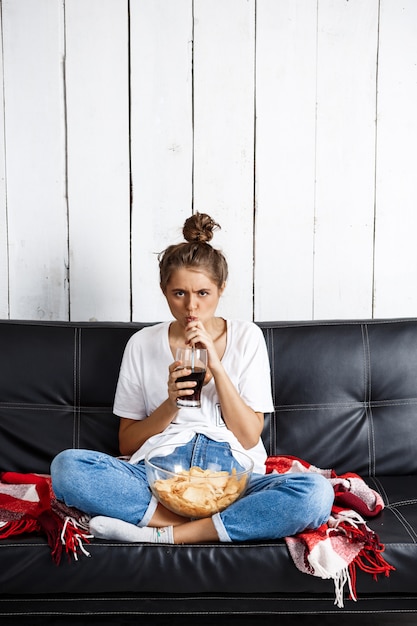 Free photo woman eating chips, drinking soda, watching tv, sitting at sofa.