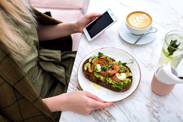 Woman eating her tasty brunch on hipster cafe, top view of marble table, salmon avocado toast , coffee and sweet tasty cheesecakes, enjoying her breakfast.