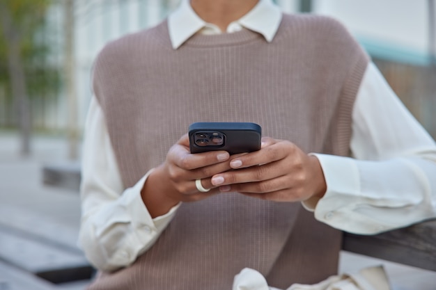 woman in elegant clothes holds mobile phone surfs wireless internet makes shopping online sits on bench outside connected to wireless internet communicates distantly