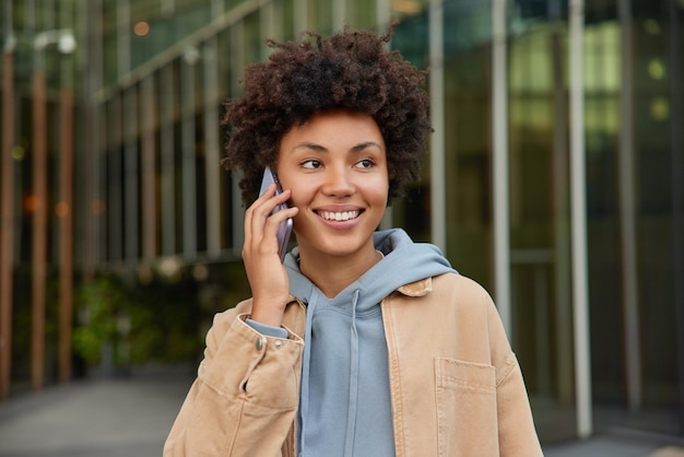 woman enjoys cellular communication smiles positively dressed casually poses outdoors makes telephone call