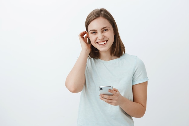 Free Photo woman exchanging numbers with cute guy on festival. charming friendly-looking young girl flicking hair behind ear flirty and laughing while gazing holding smartphone against gray wall