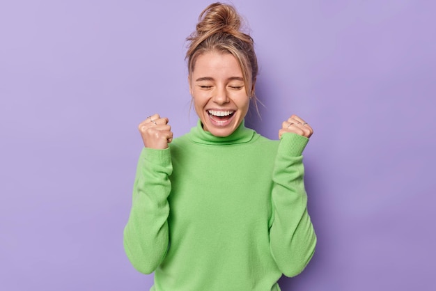 Free photo woman exclaims loudly clenches fists rejoices success feels triumph wears green turtleneck isolated on purple. onjoyed female model celebrates victory