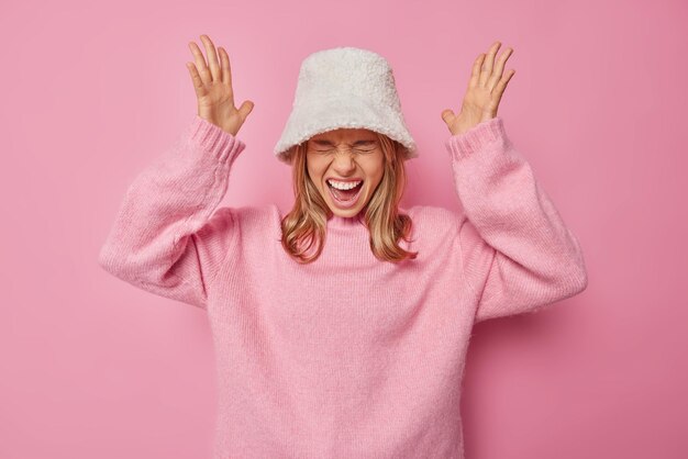 Free Photo woman exclaims loudly reacts on something awesome dressed in casual jumper and hat feels upbeat isolated on pink