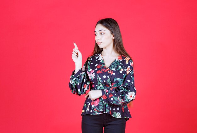 woman in floral shirt standing on red wall and showing upside. 