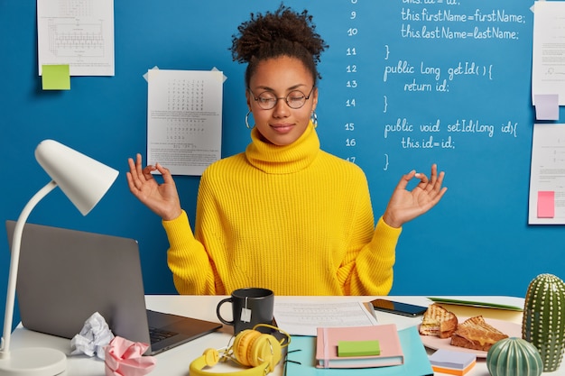 Free Photo woman freelance worker does yoga exersice at workplace, enjoys calm tranquil atmosphere, wears round glasses and jumper