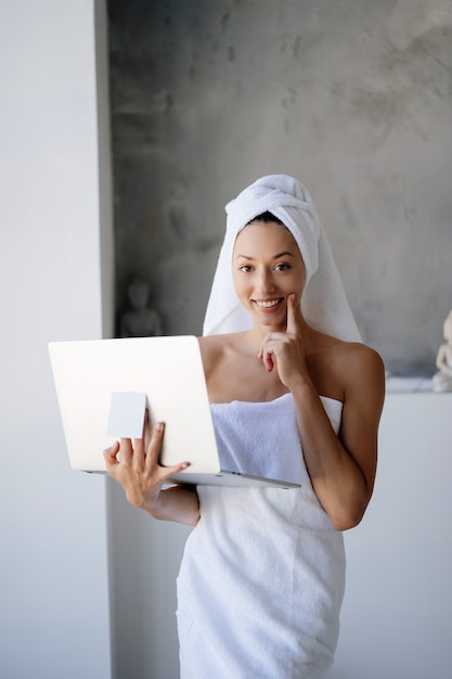 Woman freelancer in white towel stand in the bathroom with a laptop