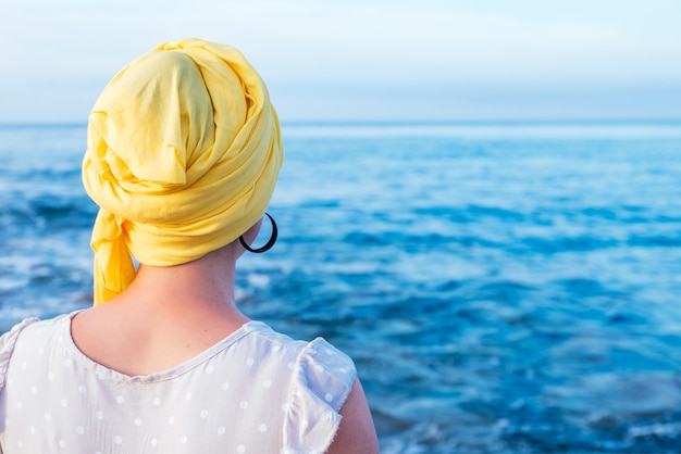 Free photo woman from behind with yellow scarf covering her head without hair contemplating the sea horizon