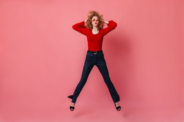 Free Photo woman in great cheerful mood is jumping on pink space. lady in glasses, red blouse and pants whistling and touching her curly hair.