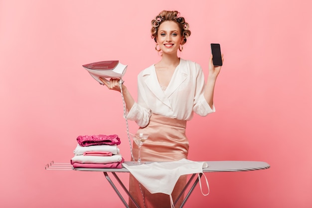 woman in great mood poses with smartphone and iron near ironing board