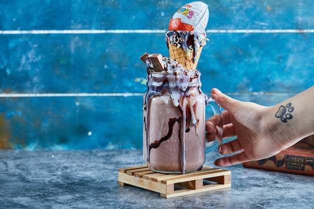 Free photo a woman hand with tattoo holding a mug of chocolate smoothie with kinder surprise, sprinkles in the blue surface