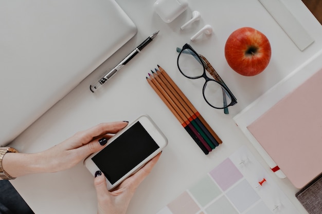 Woman hands holding smartphone at desk with pencils, glasses and laptop