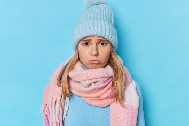 woman has gloomy expression as someone hurt her feelings complains about something wears knitted hat and scarf around neck isolated on blue feels guilty