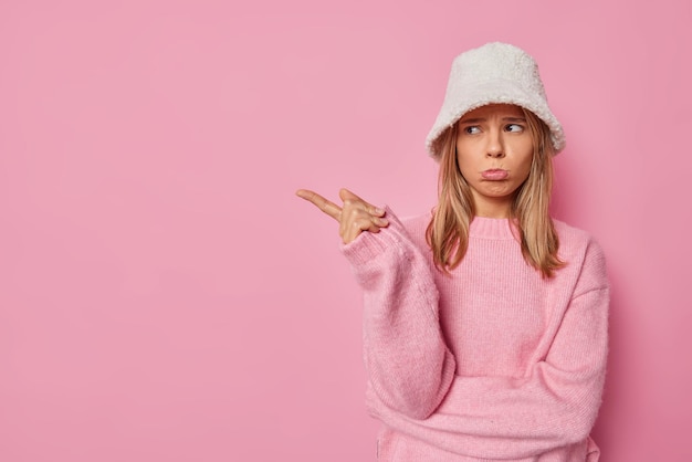 Free photo woman has sulking expression purses lips and points away with index finger wears white fur hat and jumper isolated on pink