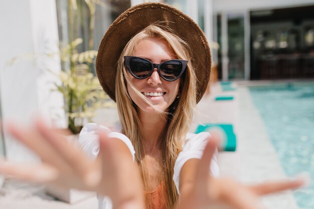 woman in hat making selfie at resort with wonderful smile. Beautiful european female tourist having fun near pool.