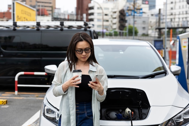 Woman having a coffee break while her electric car is charging and using smartphone