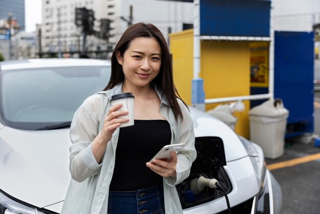 Woman having a coffee break while her electric car is charging and using smartphone