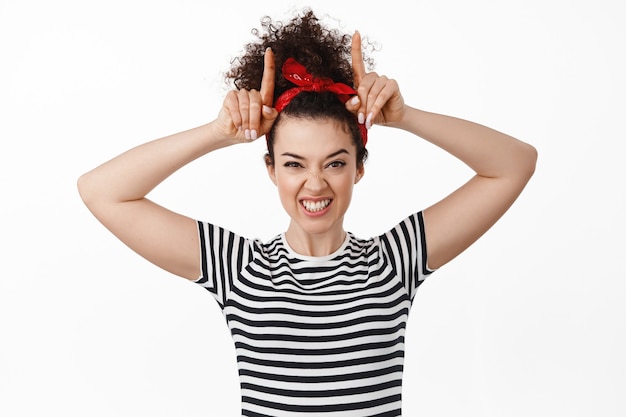 Free photo woman in headband showing tongue, bull horns gesture, looking stubborn and playful, standing on white