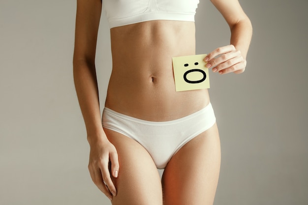 Free Photo woman health. female model holding card with emoji near stomach. young adult girl with paper for sign or symbol isolated on gray studio background. cut out part of body. medical problem and solution.