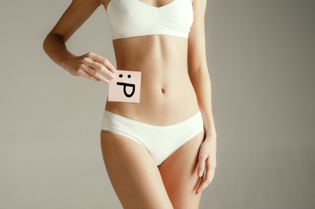 Free Photo woman health. female model holding card with emoji near stomach. young adult girl with paper for sign or symbol isolated on gray studio background. cut out part of body. medical problem and solution.