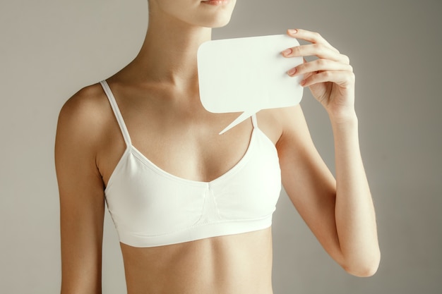 Woman health. Female model holding empty card near breast. Young adult girl with paper for sign or symbol isolated on gray studio background. Cut out part of body. Medical problem and solution.