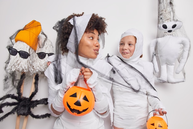  woman and her daughter pretend being ghosts holds pumpkins prepare for halloween mascarade looks gladfully at each other surrounded by handmade attributes. Autumn mystery holiday