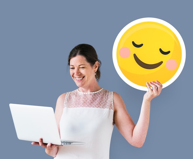 Free photo woman holding a blushing emoticon and using a laptop