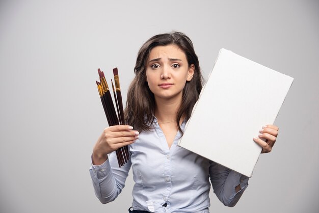 Woman holding empty canvas and paintbrushes on gray wall. 