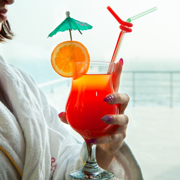 Woman holding red cocktail looking outside from window in bathrobe .