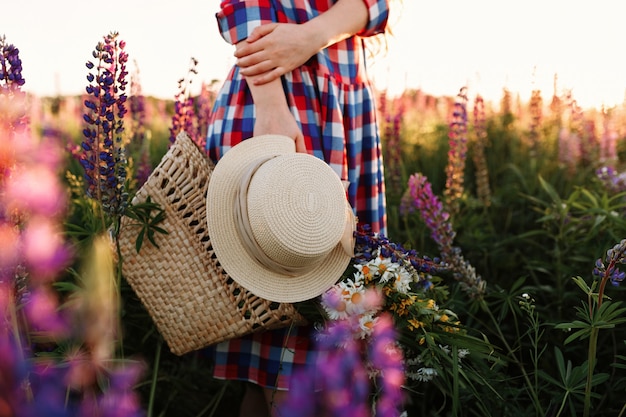 Woman holding straw bag and hat, standing in flower field at sunset. 