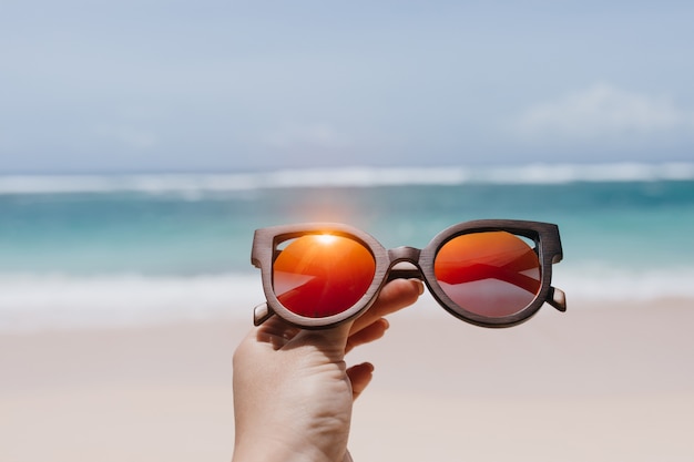 Free photo woman holding stylish summer sunglasses over sea. outdoor photo of female hand with glasses at beach.