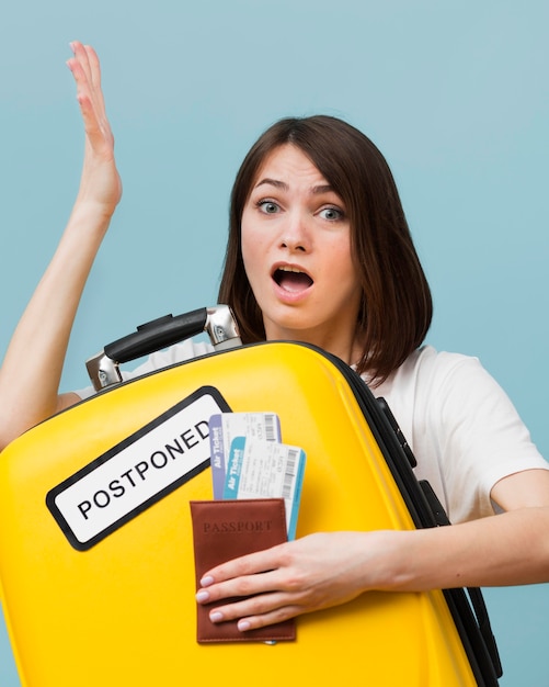 Woman holding a yellow baggage with a postponed sign while holding airplane tickets