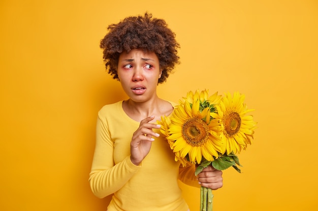 Free photo woman holds bouquet of sunflowers has red eyes wears casual jumper isolated over vivid yellow