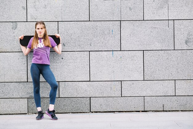 Woman holds a skateboard standing in front of a grey wall