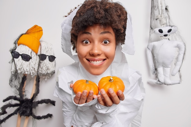 Free Photo woman holds two small pumpkins prepares decorations for halloween holiday smiles happily poses on white with creepy creatures around