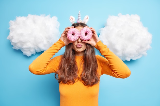 Free photo woman holds two sweet baked doughnuts on eyes keeps lips folded enjoys eating tasty high calorie food dressed in orange jumper isolated on blue