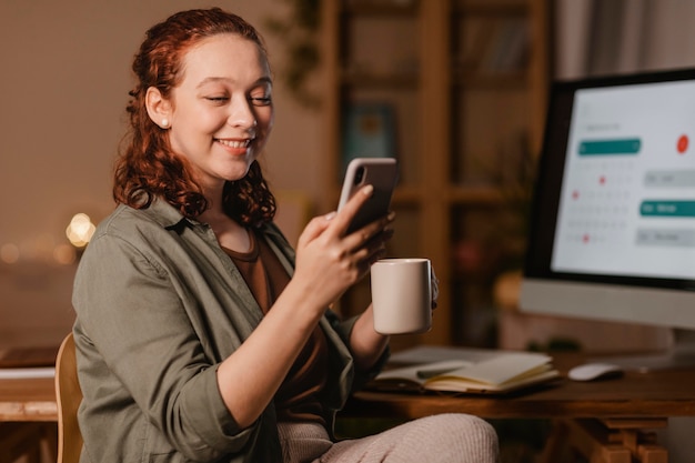 Woman at home using smartphone in front of the computer while having coffee