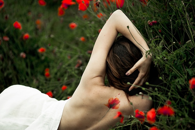 Woman is lying with naked back with a tattoo on it covered a white shirt among poppy flowers