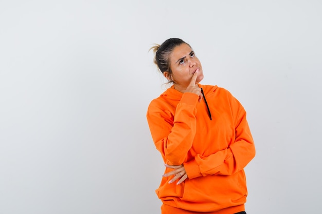 Free photo woman keeping finger on lips in orange hoodie and looking pensive