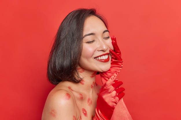 woman keeps eyes closed smiles pleasantly shows white even teeth touches face gently poses with naked shoulders on red feels happy