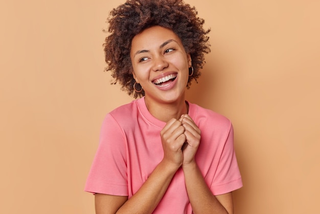 woman keeps hands together smiles broadly and looks away expresses positive emotions wears casual pink t shirt isolated on beige admires something nice.