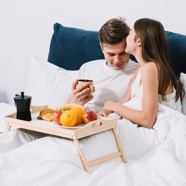 Woman kissing man on forehead on bed with food tray