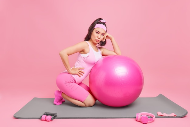 woman leans at inflated fitball has fintess training at home gym has combed pony tail wears sportsclothes leads healthy lifestyle isolated on pink 